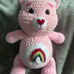 May include: A light pink crocheted teddy bear with a white muzzle, black plastic eyes, and a red heart-shaped nose. The bear features a white circular patch on its belly displaying a colorful embroidered rainbow arching over a small blue cloud.