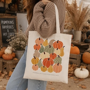 Peut inclure: Un sac fourre-tout en toile couleur crème avec une impression graphique colorée représentant diverses citrouilles et courges dans des tons orange, vert et jaune. Le texte "PUMPKIN SPICE AND EVERYTHING NICE" est imprimé en petites lettres noires.