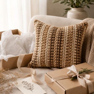 May include: A square throw cushion featuring a textured crochet pattern with vertical stripes in shades of brown, tan, and cream. The cushion has a dense, repeating stitch design and is positioned next to open gift boxes with tissue paper and ribbon.