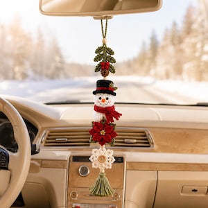 May include: A crocheted car hanging ornament featuring a green holly sprig with red berries, a white snowman wearing a black top hat and a red scarf, a red poinsettia flower, a white snowflake with a central wooden button, and a green tassel.