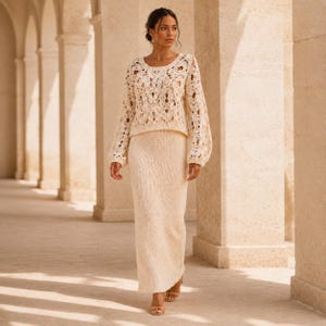 May include: A two-piece cream-colored outfit consisting of a long-sleeved, open-knit crochet sweater with a detailed floral pattern and a matching floor-length, solid-knit maxi skirt. The ensemble features a consistent light beige hue throughout.