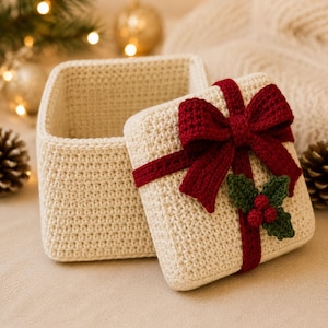 Peut inclure: Une boîte carrée crochetée blanc cassé avec un couvercle assorti. Le couvercle présente un ruban crocheté rouge foncé noué en nœud, agrémenté de deux feuilles de houx crochetées vertes et de trois petites baies rouges au centre.