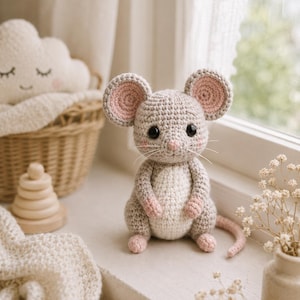 Puede incluir: Un pequeño juguete de ratón tejido a ganchillo a mano con hilo gris claro, vientre color crema y detalles en rosa suave en el interior de las orejas, las patas y la nariz. El ratón tiene orejas grandes y redondas, ojos negros y bigotes blancos.