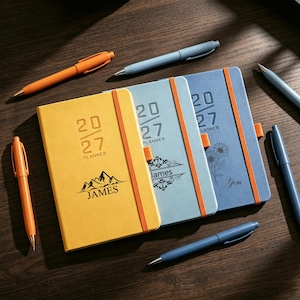 May include: Three rectangular 2027 planners with elastic bands, one yellow and two blue, featuring personalized names and graphics. The yellow planner shows a mountain design and the name James. The blue planners feature floral and scroll motifs with the names James and Gem, accompanied by several orange and blue ballpoint pens.