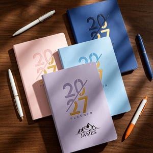 May include: Four 2027 planners in pink, light blue, lavender, and dark blue, each featuring a diagonal slash design and the word PLANNER. The lavender planner includes a mountain graphic and the name JAMES. Four pens in white, blue, and orange.