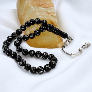 Peut inclure: Un chapelet composé de perles noires rondes et lisses ornées de petits points blancs. Le cordon comprend une perle noire plus grande et un pompon en métal argenté terminé par deux petites sphères argentées au bout de la chaînette.