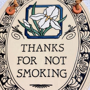 May include: An oval ceramic wall plaque with a cream background and a black decorative border. It features a central illustration of a white iris flower and the black text THANKS FOR NOT SMOKING. Two brown leather cords are attached to the top.