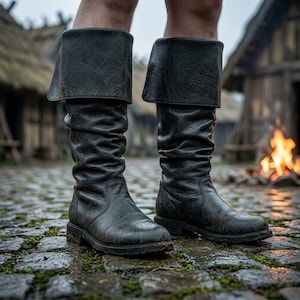 Puede incluir: Un par de botas altas de cuero negro con un borde ancho doblado en la parte superior, punta redondeada y un acabado texturizado y desgastado. Las botas tienen un diseño de caña holgada y fruncida con un tacón bajo y plano.