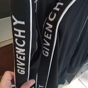 May include: A black track jacket featuring white side stripes with the text GIVENCHY embroidered in white. The garment includes a silver-toned zipper closure along the side panel, with the brand name repeated vertically along the sleeve trim.