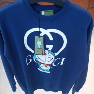 May include: A navy blue long-sleeve sweatshirt featuring a large white interlocking GG logo and a graphic of the character Doraemon. A rectangular green tag hangs from the collar, displaying the text Doraemon x Gucci and an image of Doraemon.