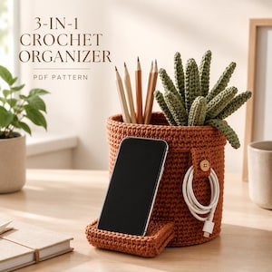 Puede incluir: Un organizador de ganchillo cilíndrico de color óxido con soporte para smartphone integrado, una pequeña pestaña con botón para cables y una suculenta de ganchillo verde. El texto 3-IN-1 CROCHET ORGANIZER PDF PATTERN es visible.