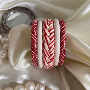 Può includere: Un set di cinque braccialetti impilati composto da due bracciali rossi con linee ondulate bianche, due bracciali color bianco sporco a tinta unita e un bracciale centrale intrecciato con un motivo a righe rosse e bianche, disposti in fila verticale.
