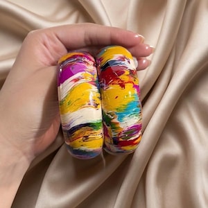 Op de afbeelding: Twee dikke armbanden met een glanzend, veelkleurig abstract verfwervelpatroon. Het levendige ontwerp bevat gedurfde strepen van geel, magenta, dieprood, felblauw, groen en wit, wat zorgt voor een dynamische en kleurrijke uitstraling.