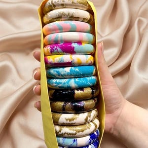 Può includere: Una pila di tredici braccialetti rotondi tenuti insieme da una custodia di carta gialla. I braccialetti presentano vari motivi marmorizzati in colori tra cui bianco, oro, rosa, blu, giallo, nero e viola, con una finitura liscia e lucida.