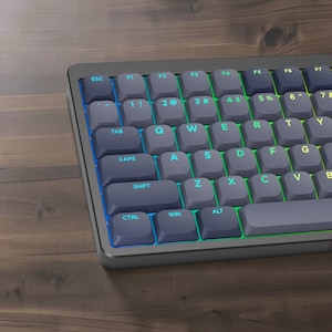 May include: A mechanical keyboard featuring dark gray rounded keycaps with light blue and green backlit lettering. The keys include standard QWERTY layout markings such as ESC, TAB, CAPS, SHIFT, CTRL, WIN, and ALT, with vibrant light emitting from beneath.