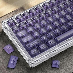 May include: A mechanical keyboard featuring a transparent frame and translucent purple keycaps with white lettering. The layout includes standard alphanumeric keys, function keys, and modifier keys. Three loose purple keycaps are positioned nearby.