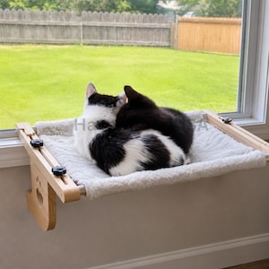 May include: A light-colored wooden cat window perch featuring a soft, white, plush fabric mat. The frame is constructed with adjustable side supports and black knobs, and the side panel includes a decorative cutout in the shape of a paw print.