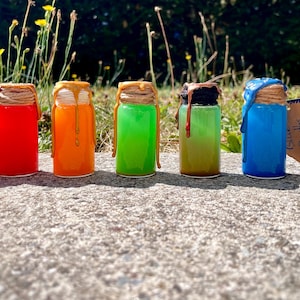 Könnte beinhalten: Sieben kleine Glasflaschen gefüllt mit leuchtenden Flüssigkeiten in Pink, Rot, Orange, Grün, Gelbgrün, Blau und Schwarz. Jede Flasche hat einen mit Schnur umwickelten Korken, an dem farbiges Wachs herabläuft. Ein kleines braunes Etikett trägt die Aufschrift Blue Depths.