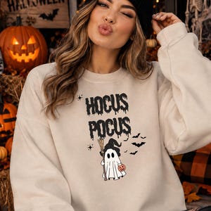 Könnte beinhalten: Ein beigefarbenes Sweatshirt mit Rundhalsausschnitt und einem schwarzen Grafikdruck, der HOCUS POCUS in einer tropfenden Schriftart zeigt. Unter dem Text befindet sich ein weißes Gespenst mit schwarzem Hexenhut, das einen Besen und einen Kürbis hält.