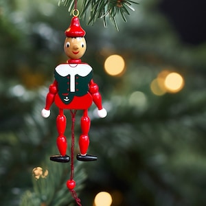 May include: A wooden marionette ornament featuring a painted figure with a red hat, a dark green vest with white accents, and red limbs. The figure has a round face with black eyes and a red smile, suspended by a string with a small red bead at the base.