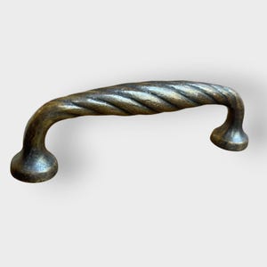 May include: A bronze-colored cabinet pull with a twisted rope design. The handle curves upward with rounded ends, creating a decorative and functional hardware piece. The pull is designed for easy gripping and adds a vintage touch.