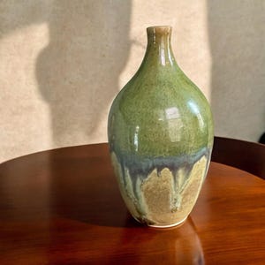 May include: A ceramic vase with a narrow neck and a rounded body, featuring a glossy glaze that transitions from a moss green at the top to a muted blue-grey band, ending in a textured, sandy beige base with visible vertical drip patterns.