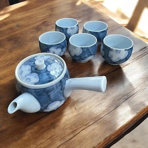 May include: A ceramic tea set with a blue and white floral design. The set includes a teapot with a handle and lid, and six matching teacups. The tea set is placed on a wooden surface.