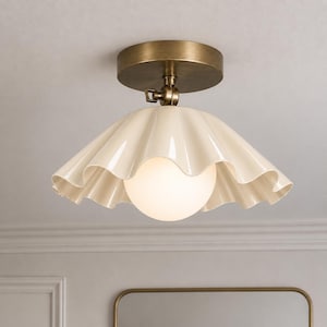 Puede incluir: Una lámpara de techo con una base circular de latón antiguo y una articulación giratoria central. La lámpara incluye una pantalla de cerámica ondulada de color crema que rodea una bombilla esférica de cristal blanco esmerilado.