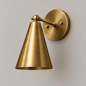 May include: A wall-mounted sconce light fitting featuring a brushed brass finish. The design includes a circular wall plate with two small mounting knobs and a conical shade attached to a short, adjustable arm with a spherical pivot joint.