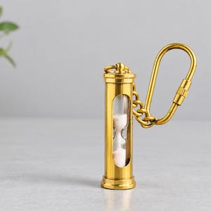 Könnte beinhalten: Ein goldfarbener Sanduhr-Schlüsselanhänger mit einem zylindrischen Rahmen und einem transparenten Glaseinsatz, der weißen Sand enthält. Die Sanduhr ist über eine kurze Metallkette mit einem passenden goldfarbenen Karabinerhaken verbunden.