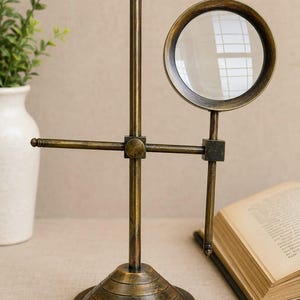 May include: A brass-toned metal magnifying glass mounted on an adjustable stand. The device features a circular lens frame attached to a horizontal arm, which is connected to a vertical post supported by a tiered, circular base with a dark finish.