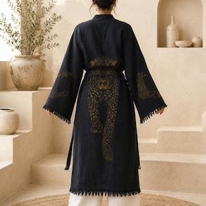 Op de afbeelding: Een donkerblauwe kamerjas met lange mouwen en een gouden luipaardprint op de rug en mouwen. Het kledingstuk heeft een ceintuur, wijde mouwen en een gefranjerde zoom, gedragen over een losvallende witte broek voor een gelaagde uitstraling.