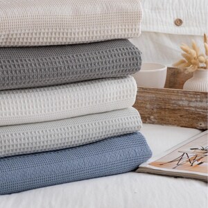 Puede incluir: Una pila de cinco mantas dobladas con un patrón de tejido de gofre texturizado. Las mantas son de colores beige, gris oscuro, blanco, gris claro y azul polvoriento, dispuestas en una pila vertical con detalles de cuadrícula visibles.