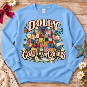 May include: A light blue crewneck sweatshirt featuring a graphic of a patchwork coat with the text DOLLY COAT OF MANY COLORS TENNESSEE. The design includes colorful quilt patterns, butterflies, daisies, stars, and a red heart on the coat.
