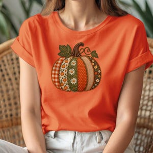 Könnte beinhalten: Ein leuchtend orangefarbenes T-Shirt mit kurzen Ärmeln und einer großen Kürbisgrafik in der Mitte. Der Kürbis ist in vertikale Segmente unterteilt, die verschiedene Muster wie orangefarbenes Vichy-Karo, weiße Gänseblümchen auf Grün und orangefarbene Punkte aufweisen.