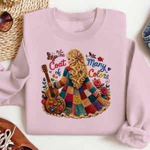 May include: A light pink crewneck sweatshirt featuring a graphic illustration of a person with long blonde hair wearing a multi-coloured patchwork coat. The person holds an acoustic guitar, surrounded by flowers and a butterfly with the text The Coat of Many Colors.