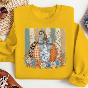 May include: A mustard yellow crewneck sweatshirt featuring a central graphic of a pumpkin with varied segments including leopard print, orange plaid, blue floral, and orange polka dots, topped with a blue bow and surrounded by colourful flowers.