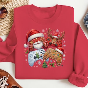 Op de afbeelding: Een rood sweatshirt met ronde hals en een feestelijke afbeelding van drie videogamecontrollers. Eén controller draagt een kerstmuts en baard, een andere heeft rendiergeweien en de derde is een peperkoek-stijl controller met een kerstmuts.