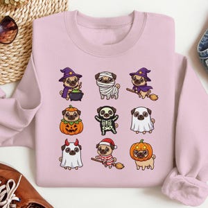 May include: A light pink crewneck sweatshirt featuring a graphic print of nine cartoon pugs dressed in various Halloween costumes, including a witch, a mummy, a pumpkin, a skeleton, a ghost, a devil, and a pug riding a broomstick.