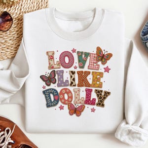 May include: A white crewneck sweatshirt featuring the text LOVE LIKE DOLLY in large, multi-patterned block letters. The design includes various textures like floral, leopard print, polka dots, and gingham, accented by several colorful butterflies.
