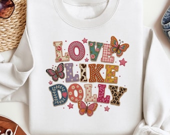 May include: A white crewneck sweatshirt featuring the text LOVE LIKE DOLLY in large, multi-patterned block letters. The design includes various textures like floral, leopard print, polka dots, and gingham, accented by several colorful butterflies.