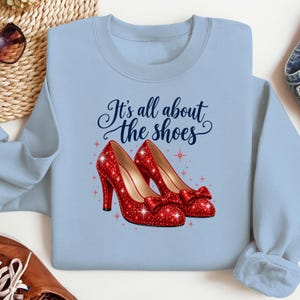 Pode incluir: Uma camisola de gola redonda azul-clara com um gráfico de dois sapatos de salto alto vermelhos com purpurinas e laços. Acima dos sapatos, o texto "It's all about the shoes" está escrito em azul-escuro, rodeado por pequenos detalhes de estrelas.