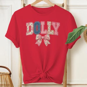 Könnte beinhalten: Ein leuchtend rotes Kurzarm-T-Shirt mit dem Wort DOLLY, das in einem Patchwork-Stil mit Blumen- und Vichy-Karomustern gedruckt ist. Unter dem Text befindet sich eine große, passende Schleifengrafik. Das T-Shirt ist vorne unten verknotet.