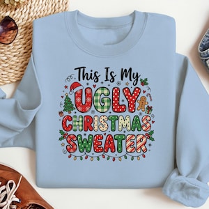 Puede incluir: Una sudadera azul claro de cuello redondo con un estampado gráfico que dice This Is My UGLY CHRISTMAS SWEATER en letras rojas, verdes y blancas. El diseño incluye iconos festivos como un gorro de Papá Noel y un árbol de Navidad.