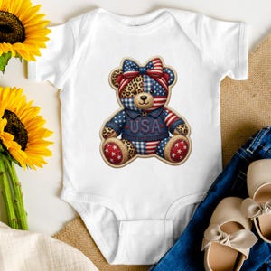 Op de afbeelding: Een wit babyrompertje met korte mouwen en een centrale afbeelding van een teddybeer die een spijkerjasje draagt met de tekst USA. De beer heeft een patchworkontwerp met luipaardprint, blauw-witte ruiten en rood-witte strepen.
