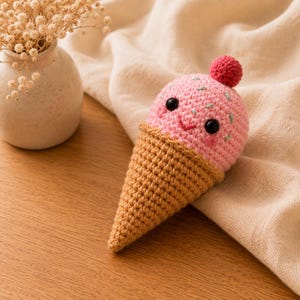 Könnte beinhalten: Ein gehäkeltes Eiswaffel-Spielzeug mit einer rosafarbenen Kugel, einer kleinen roten Kirsche obenauf, schwarzen Sicherheitsaugen, einem gestickten Lächeln und hellgrünen Streuseln. Die Waffel besteht aus beigem Garn mit einem strukturierten Rippenmuster.