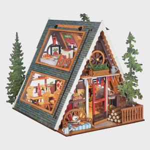 May include: A miniature A-frame cabin model featuring a green shingled roof, two levels with visible interior rooms, a red front door, a wooden porch with a bench, stacked logs, and two decorative evergreen tree cutouts on the sides.