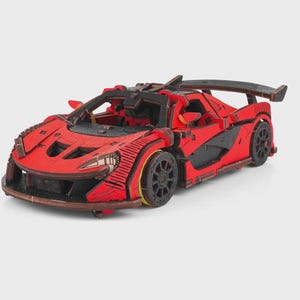 May include: A red and black wooden model sports car featuring a low-profile aerodynamic body, a large rear spoiler, intricate paneling, and detailed wheel rims. The construction displays visible layered wood edges and precise cutouts throughout the frame.