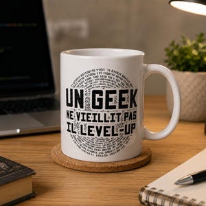 Op de afbeelding: Een witte keramische mok met zwarte tekst die UN GEEK NE VIEILLIT PAS IL LEVEL-UP leest, omringd door een cirkelvormig patroon van binaire codecijfers. De mok heeft een standaard handvat en staat op een ronde kurken onderzetter op een houten bureau.