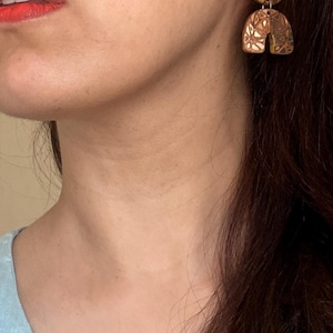 Peut inclure: Une paire de boucles d'oreilles pendantes composée d'un clou d'oreille circulaire et d'une pièce en forme d'arche semi-circulaire. Les deux éléments sont en matériau marron avec un motif géométrique sculpté de lignes et de triangles.